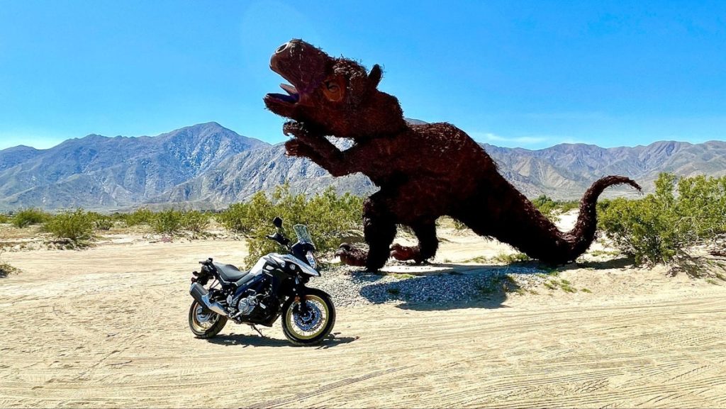Distant shot of the V-strom parked on sand next to a sand road that is next to metal dinosaur in desert landscape
